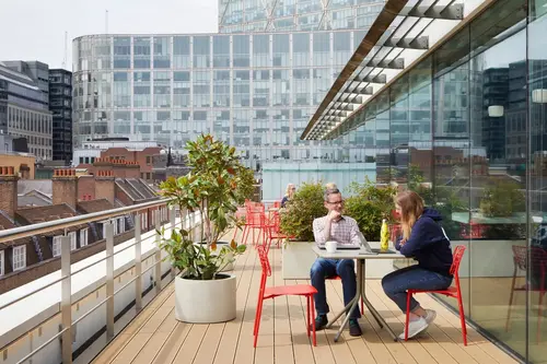 FORA Spitalfields roof terrace with four people sitting on red chairs next to green potted plants with London skylight in the background