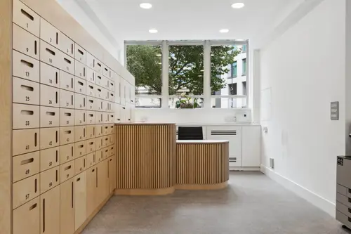 Reception area at Britannia House, 11 Glenthorne Road, with a curved slatted timber desk, a wall of numbered plywood lockers and a window overlooking trees.