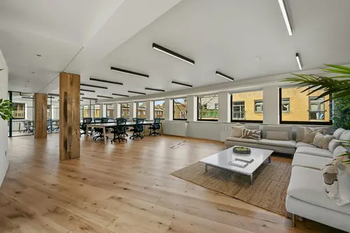 Office space at 4 7 Manchester Street showcasing two rows of wooden desks with black mesh office chairs.  There is also a breakout space with a beige L-shaped couch
