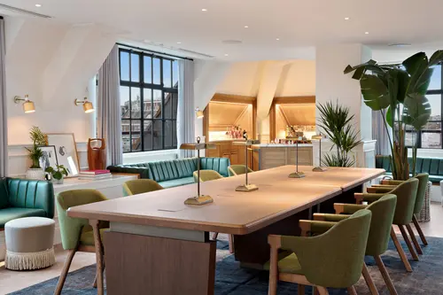 Office Space at Mortimer House in London Showcasing a long wooden desk with eight green chairs. There are four gold lamps on the table. The room has large windows and green leather bay window couches.