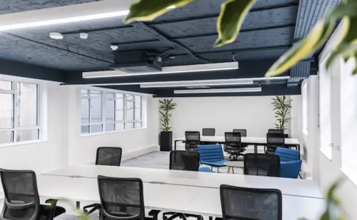 Open-plan office at 20 Ironmonger Lane with rows of white desks, black mesh chairs, blue armchairs, exposed painted ceiling services and windows along two walls.