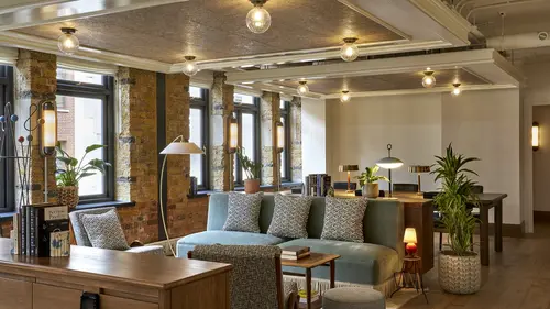 Office Space and breakout area at 1 Warwick in London. There is a blue couch with monochrome cushions, a large white standing lamp and a long desk with black office chairs. The room has an exposed brick accent wall and ambient lighting with large black paned windows.